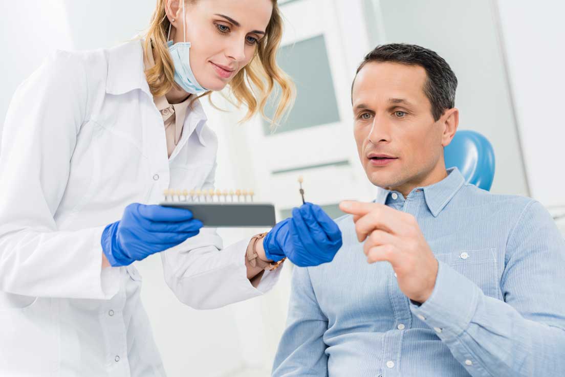 A dentist wearing gloves and a mask shows a male patient a set of tooth color samples while discussing dental implant options in a clinic.