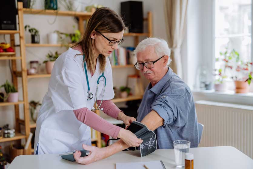A nurse checks the blood pressure of an elderly man at home, using a blood pressure monitor, while both are seated at a table.