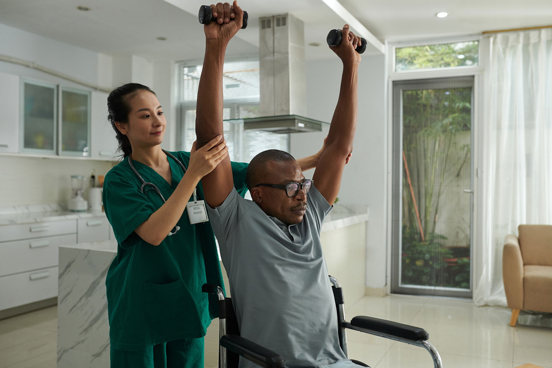 A healthcare professional assists a man in a wheelchair with physical therapy exercises. The man is lifting dumbbells while the caregiver supports his arms.