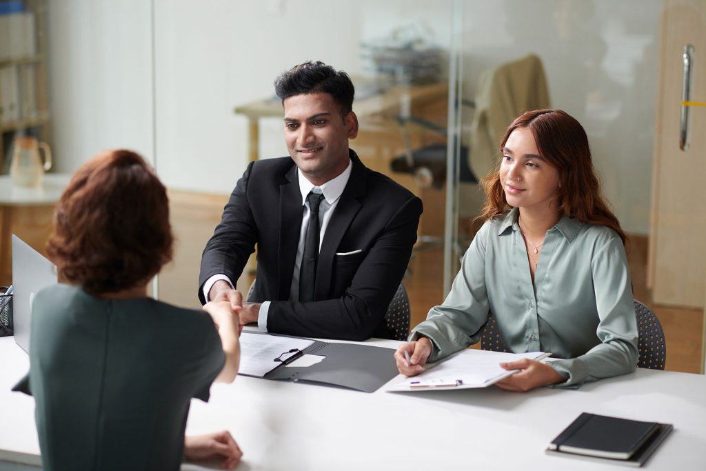 A man in a black suit shakes hands with a job candidate across the table, while a woman in a green blouse takes notes during an interview in a modern office.